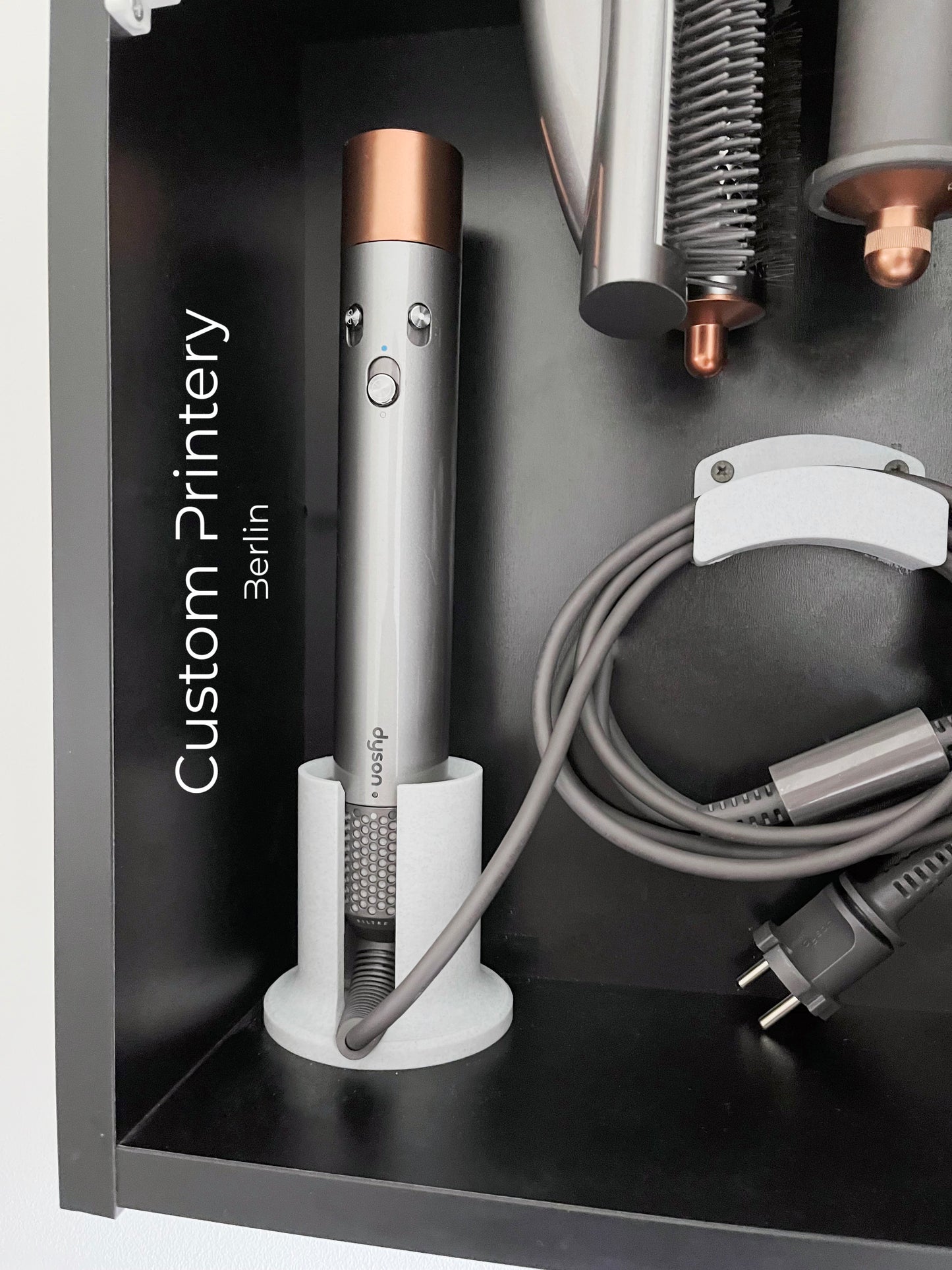 Schwarze Wandhalterung für Dyson Haarstyling-Gerät und Zubehör. Die Halterung enthält den Haartrockner, verschiedene Styling-Aufsätze und ist sauber organisiert. Oben auf dem Regal steht eine moderne Vase mit getrockneten Eukalyptuszweigen.