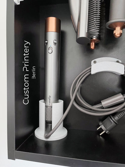 Schwarze Wandhalterung für Dyson Haarstyling-Gerät und Zubehör. Die Halterung enthält den Haartrockner, verschiedene Styling-Aufsätze und ist sauber organisiert. Oben auf dem Regal steht eine moderne Vase mit getrockneten Eukalyptuszweigen.