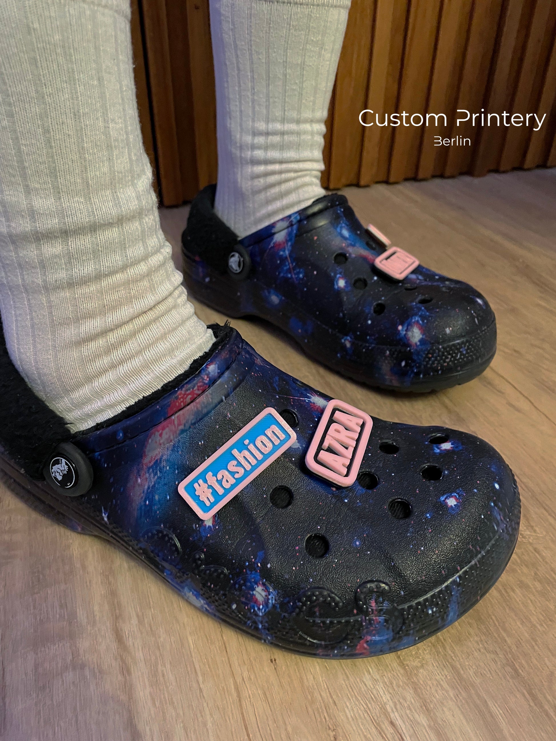 Custom galaxy-patterned Crocs with personalized name charms 'AZRA' and '#fashion' from Custom Printery Berlin, worn with white socks in an indoor setting