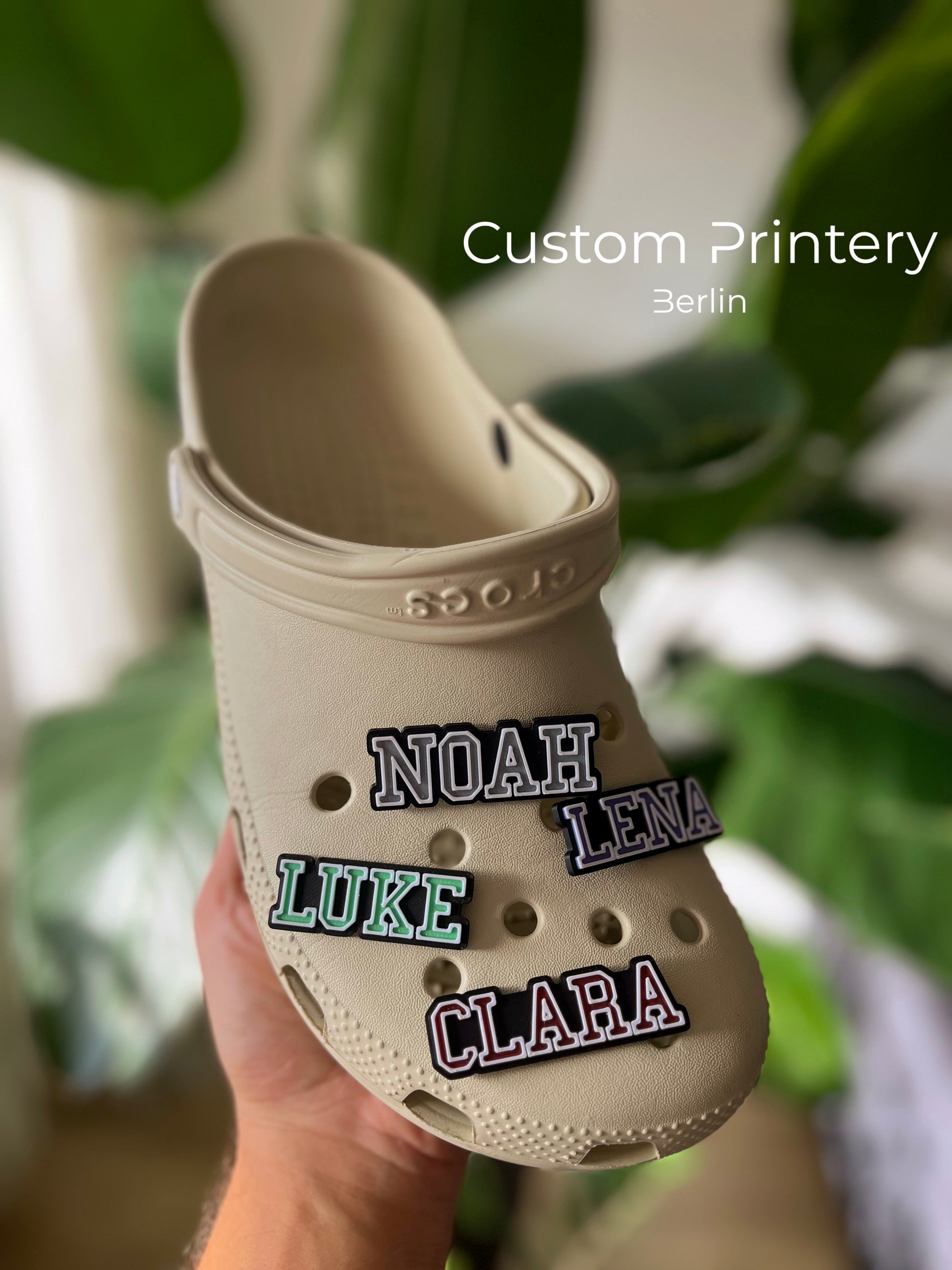 Custom black Crocs with colorful personalized name charms for 'Lena,' 'Noah,' 'Theo,' and 'Clara' from Custom Printery Berlin, showcased in a casual indoor setting with green plants.