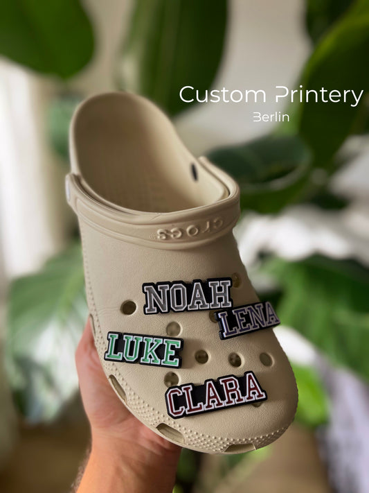 Custom black Crocs with colorful personalized name charms for 'Lena,' 'Noah,' 'Theo,' and 'Clara' from Custom Printery Berlin, showcased in a casual indoor setting with green plants.