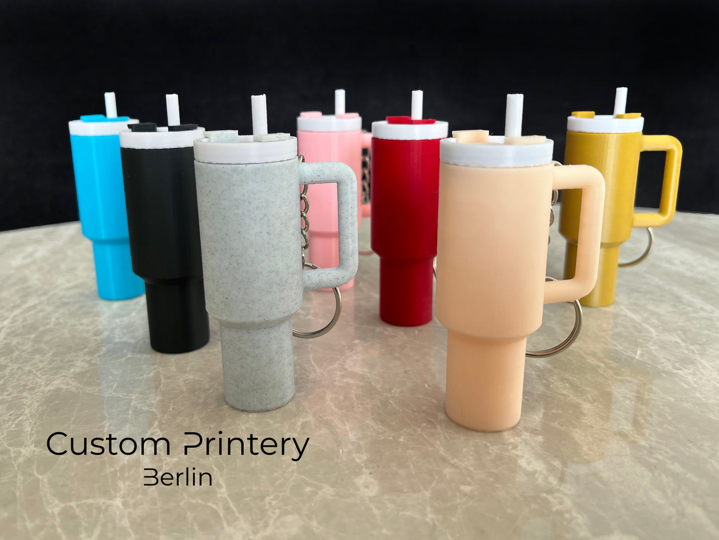 Mehrere bunte Schlüsselanhänger im Stil des Stanley Quencher Cups sind auf einer Marmoroberfläche arrangiert. Die Miniaturbecher haben Henkel, Trinkhalme und verschiedene Farben, darunter Schwarz, Blau, Pink, Rot, Gelb, Beige und Grau.