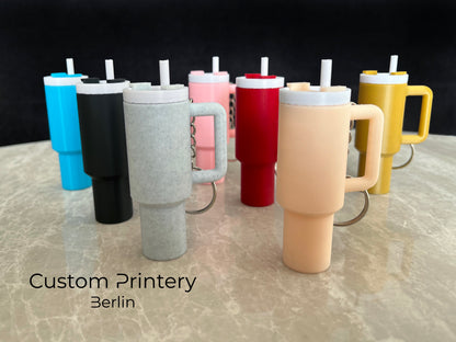 Mehrere bunte Schlüsselanhänger im Stil des Stanley Quencher Cups sind auf einer Marmoroberfläche arrangiert. Die Miniaturbecher haben Henkel, Trinkhalme und verschiedene Farben, darunter Schwarz, Blau, Pink, Rot, Gelb, Beige und Grau.