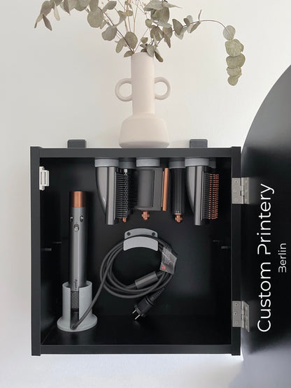Schwarze Wandhalterung für Dyson Haarstyling-Gerät und Zubehör. Die Halterung enthält den Haartrockner, verschiedene Styling-Aufsätze und ist sauber organisiert. Oben auf dem Regal steht eine moderne Vase mit getrockneten Eukalyptuszweigen.