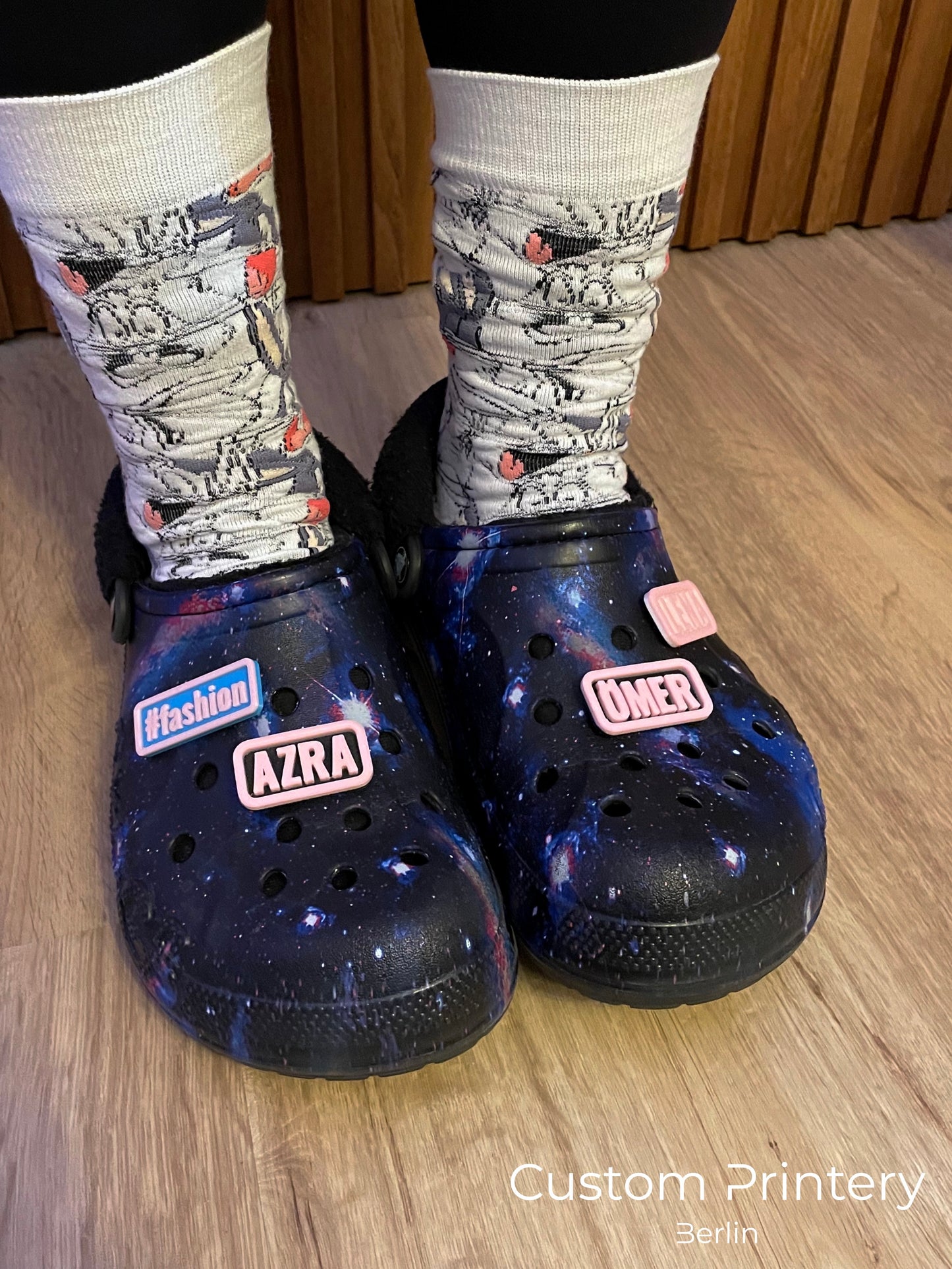 Custom galaxy-patterned Crocs with personalized name charms 'AZRA' and '#fashion' from Custom Printery Berlin, worn with white socks in an indoor setting