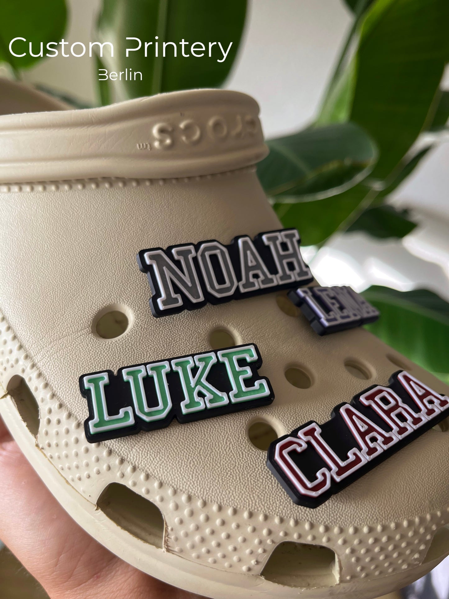 Custom black Crocs with colorful personalized name charms for 'Lena,' 'Noah,' 'Theo,' and 'Clara' from Custom Printery Berlin, showcased in a casual indoor setting with green plants.