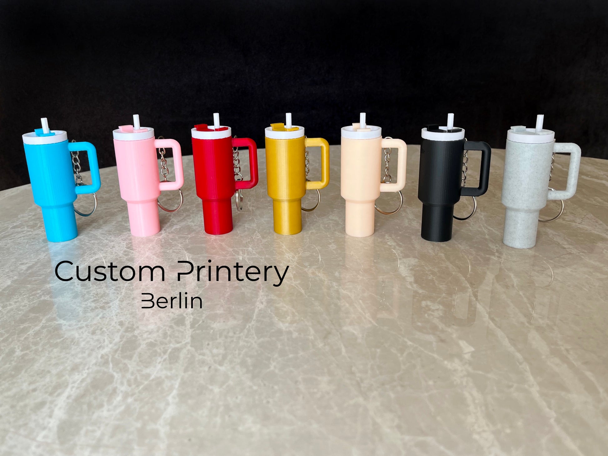 Mehrere bunte Schlüsselanhänger im Stil des Stanley Quencher Cups sind auf einer Marmoroberfläche arrangiert. Die Miniaturbecher haben Henkel, Trinkhalme und verschiedene Farben, darunter Schwarz, Blau, Pink, Rot, Gelb, Beige und Grau.