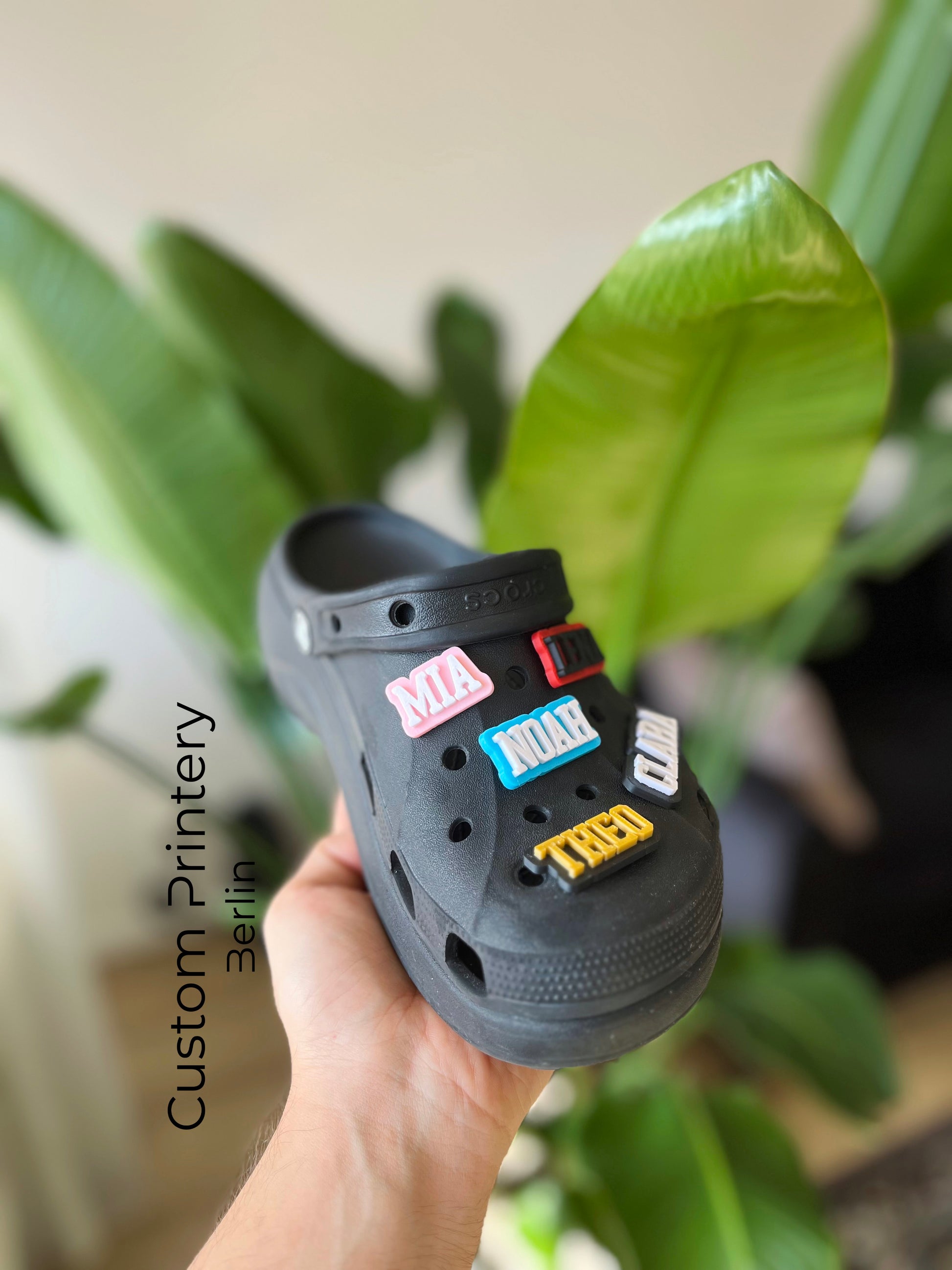 Custom black Crocs with colorful personalized name charms for 'Lena,' 'Noah,' 'Theo,' and 'Clara' from Custom Printery Berlin, showcased in a casual indoor setting with green plants.