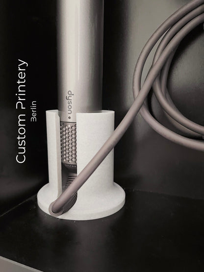 Schwarze Wandhalterung für Dyson Haarstyling-Gerät und Zubehör. Die Halterung enthält den Haartrockner, verschiedene Styling-Aufsätze und ist sauber organisiert. Oben auf dem Regal steht eine moderne Vase mit getrockneten Eukalyptuszweigen.