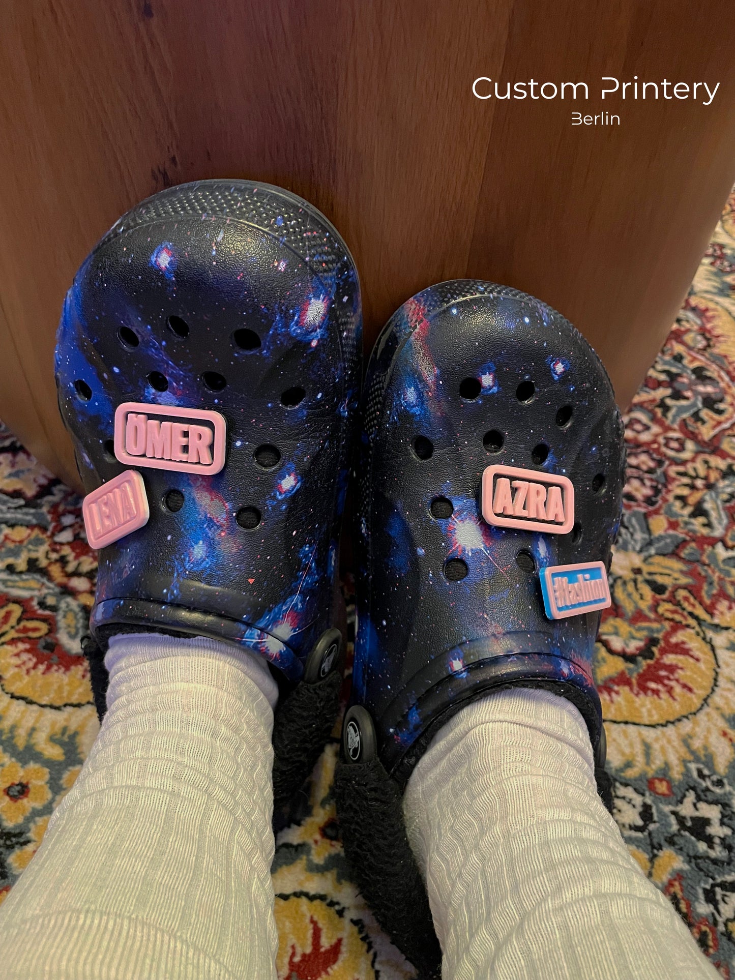 Custom galaxy-patterned Crocs with personalized name charms 'AZRA' and '#fashion' from Custom Printery Berlin, worn with white socks in an indoor setting