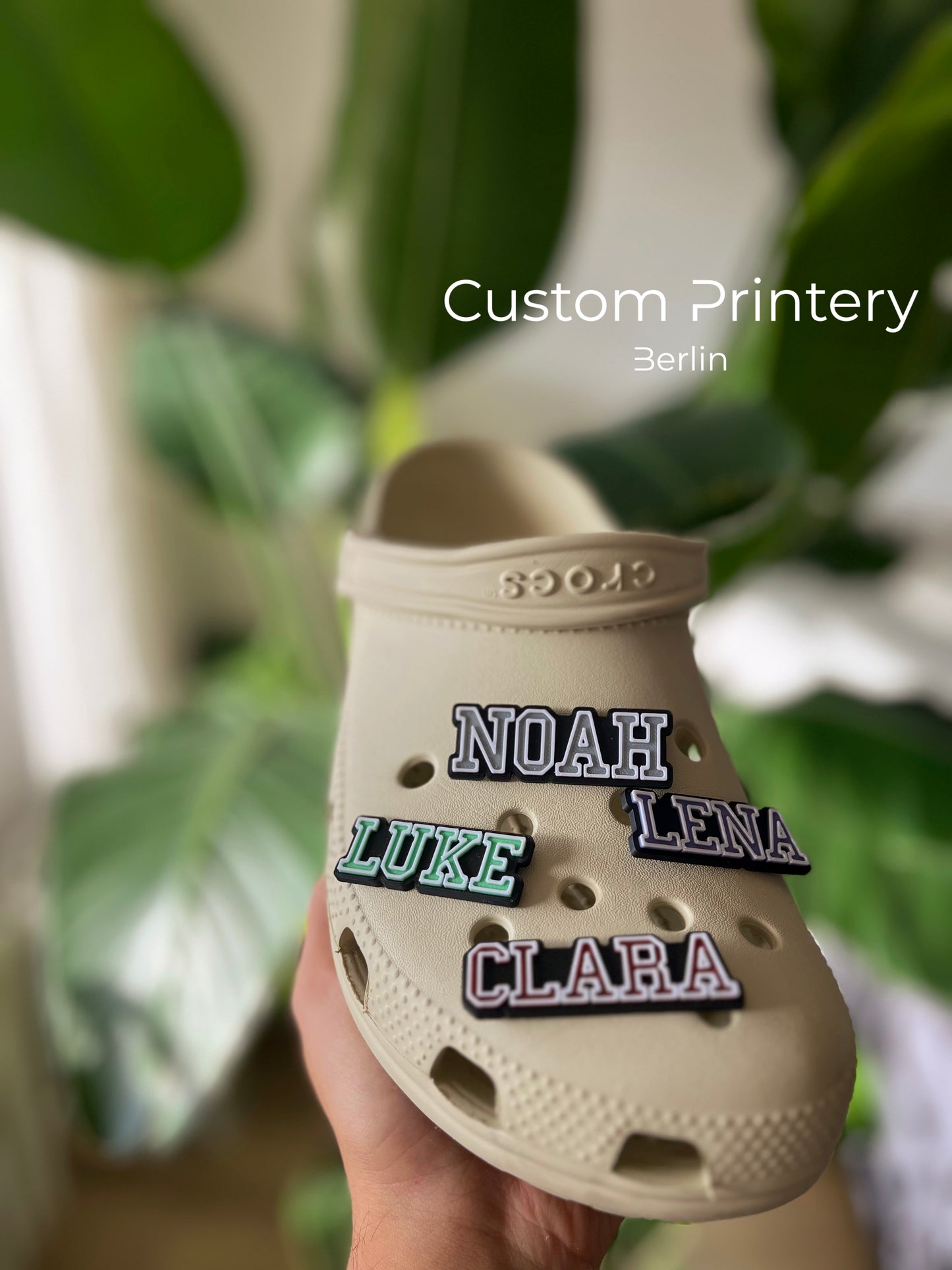 Custom black Crocs with colorful personalized name charms for 'Lena,' 'Noah,' 'Theo,' and 'Clara' from Custom Printery Berlin, showcased in a casual indoor setting with green plants.