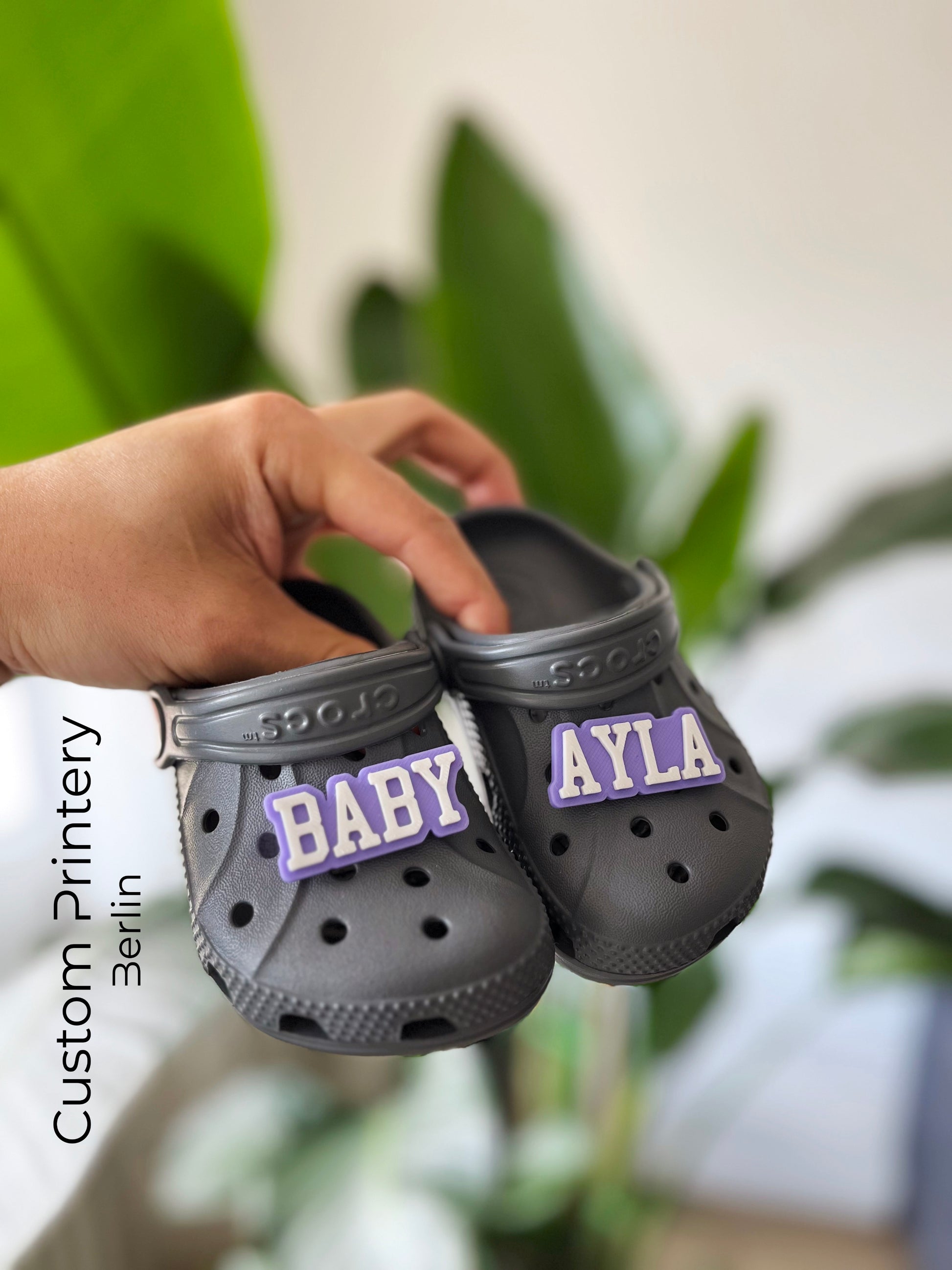 Custom black Crocs with colorful personalized name charms for 'Lena,' 'Noah,' 'Theo,' and 'Clara' from Custom Printery Berlin, showcased in a casual indoor setting with green plants.
