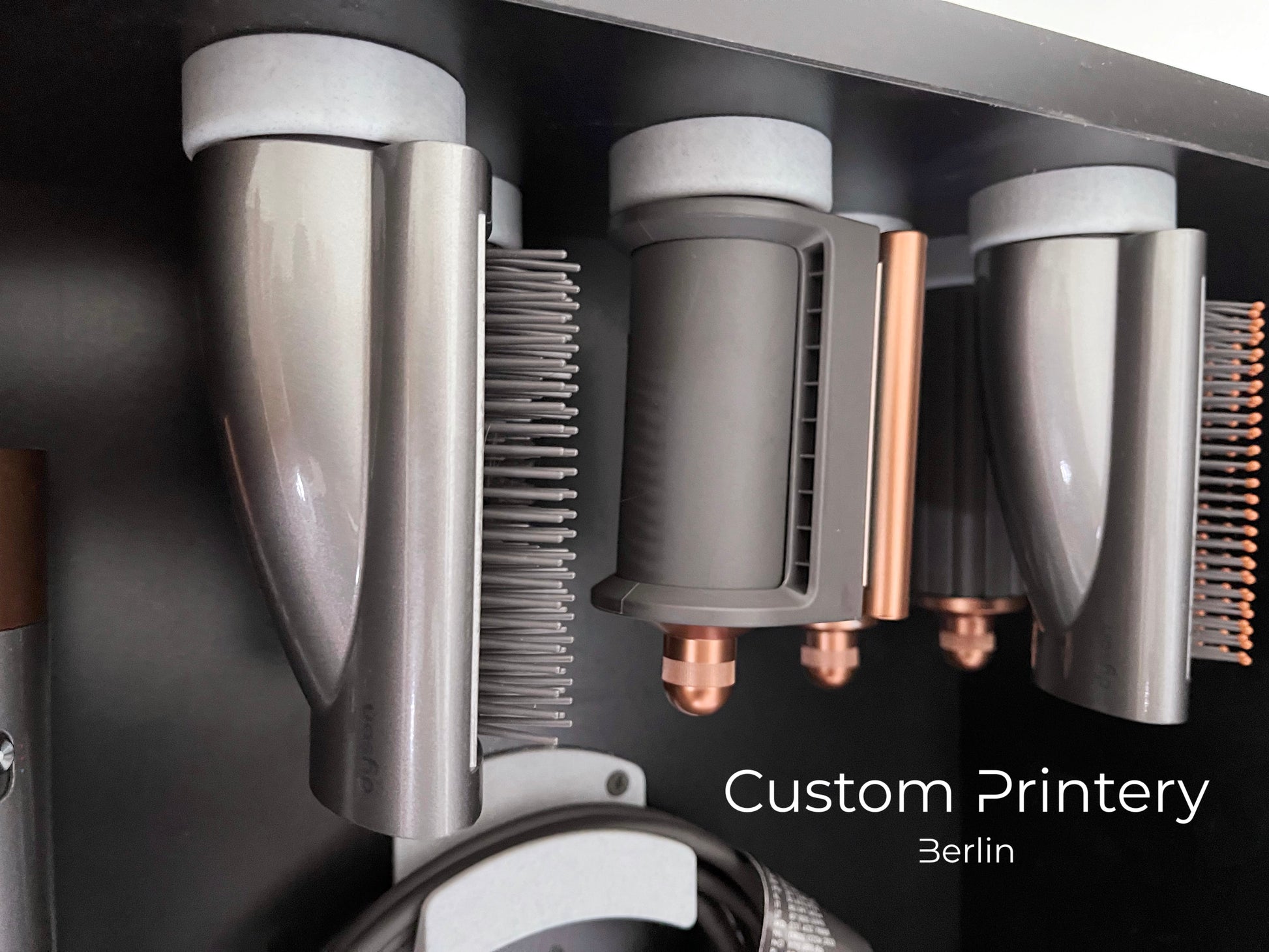 Schwarze Wandhalterung für Dyson Haarstyling-Gerät und Zubehör. Die Halterung enthält den Haartrockner, verschiedene Styling-Aufsätze und ist sauber organisiert. Oben auf dem Regal steht eine moderne Vase mit getrockneten Eukalyptuszweigen.