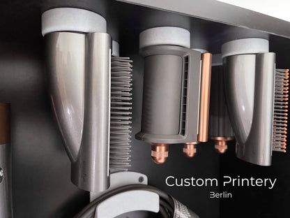 Schwarze Wandhalterung für Dyson Haarstyling-Gerät und Zubehör. Die Halterung enthält den Haartrockner, verschiedene Styling-Aufsätze und ist sauber organisiert. Oben auf dem Regal steht eine moderne Vase mit getrockneten Eukalyptuszweigen.