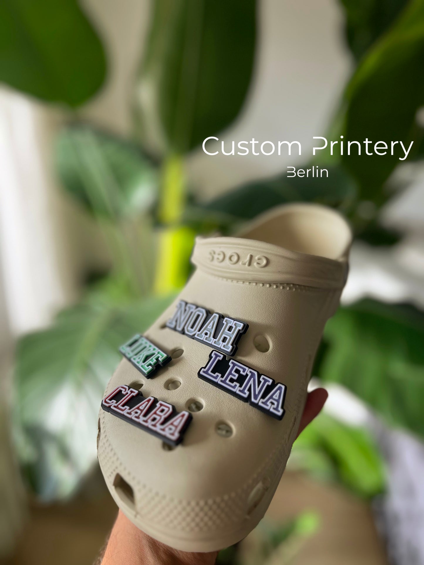 Custom black Crocs with colorful personalized name charms for 'Lena,' 'Noah,' 'Theo,' and 'Clara' from Custom Printery Berlin, showcased in a casual indoor setting with green plants.