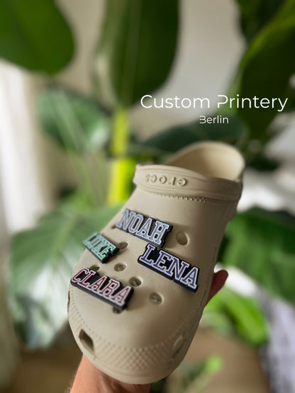Custom black Crocs with colorful personalized name charms for 'Lena,' 'Noah,' 'Theo,' and 'Clara' from Custom Printery Berlin, showcased in a casual indoor setting with green plants.