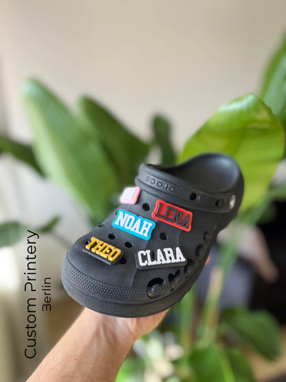 Custom black Crocs with colorful personalized name charms for 'Lena,' 'Noah,' 'Theo,' and 'Clara' from Custom Printery Berlin, showcased in a casual indoor setting with green plants.