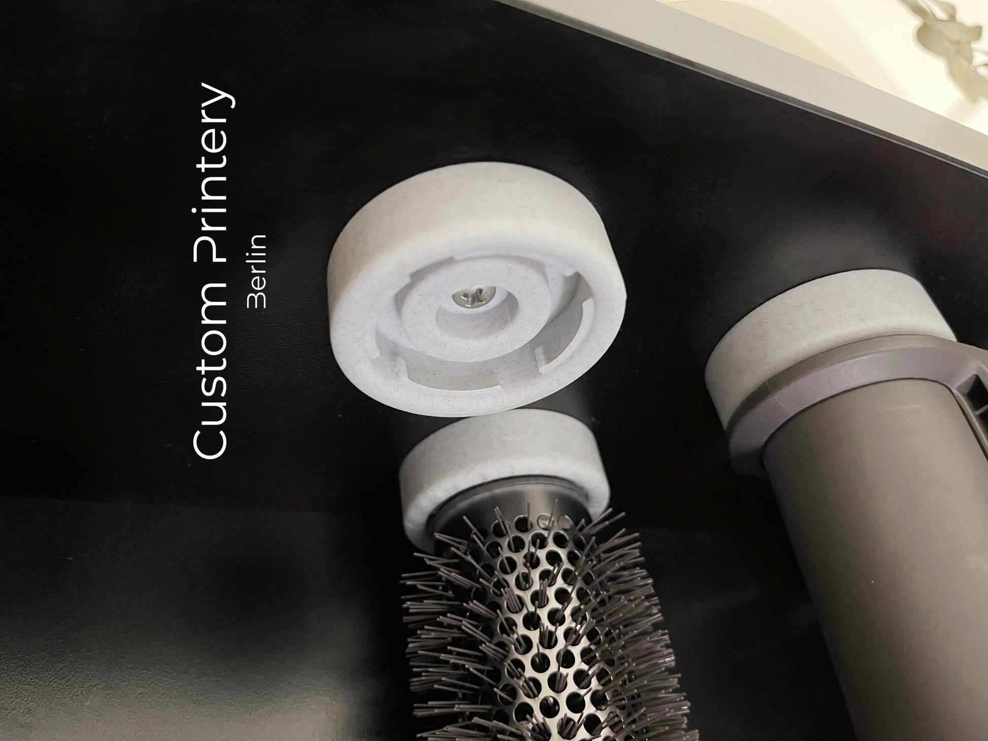 Schwarze Wandhalterung für Dyson Haarstyling-Gerät und Zubehör. Die Halterung enthält den Haartrockner, verschiedene Styling-Aufsätze und ist sauber organisiert. Oben auf dem Regal steht eine moderne Vase mit getrockneten Eukalyptuszweigen.