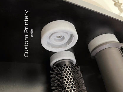 Schwarze Wandhalterung für Dyson Haarstyling-Gerät und Zubehör. Die Halterung enthält den Haartrockner, verschiedene Styling-Aufsätze und ist sauber organisiert. Oben auf dem Regal steht eine moderne Vase mit getrockneten Eukalyptuszweigen.