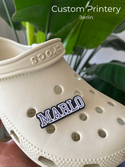 Custom black Crocs with colorful personalized name charms for 'Lena,' 'Noah,' 'Theo,' and 'Clara' from Custom Printery Berlin, showcased in a casual indoor setting with green plants.