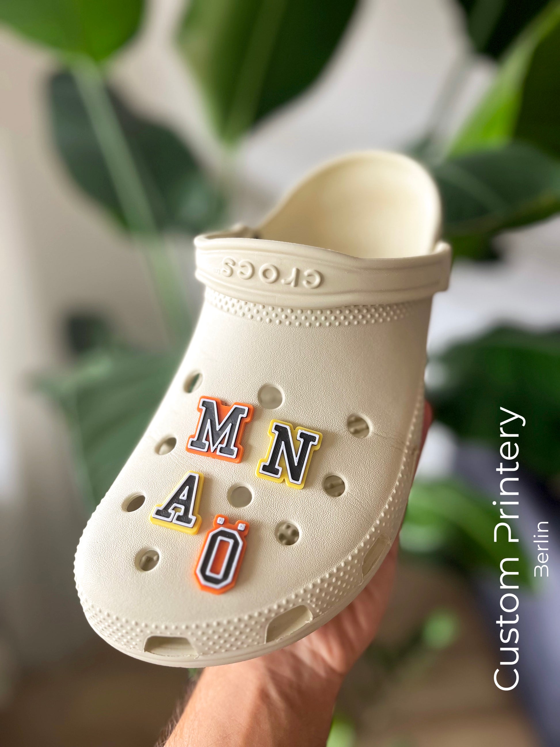 Custom black Crocs with colorful personalized name charms for 'Lena,' 'Noah,' 'Theo,' and 'Clara' from Custom Printery Berlin, showcased in a casual indoor setting with green plants.