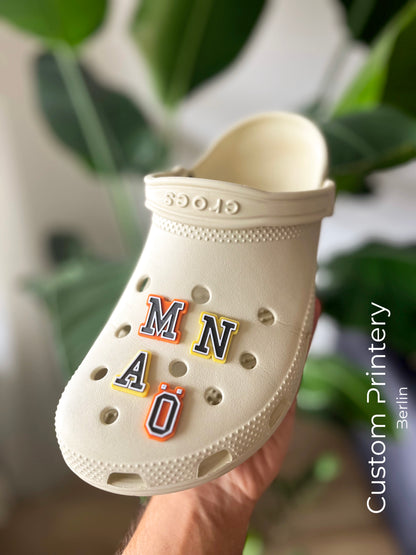 Custom black Crocs with colorful personalized name charms for 'Lena,' 'Noah,' 'Theo,' and 'Clara' from Custom Printery Berlin, showcased in a casual indoor setting with green plants.