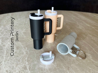 Mehrere bunte Schlüsselanhänger im Stil des Stanley Quencher Cups sind auf einer Marmoroberfläche arrangiert. Die Miniaturbecher haben Henkel, Trinkhalme und verschiedene Farben, darunter Schwarz, Blau, Pink, Rot, Gelb, Beige und Grau.