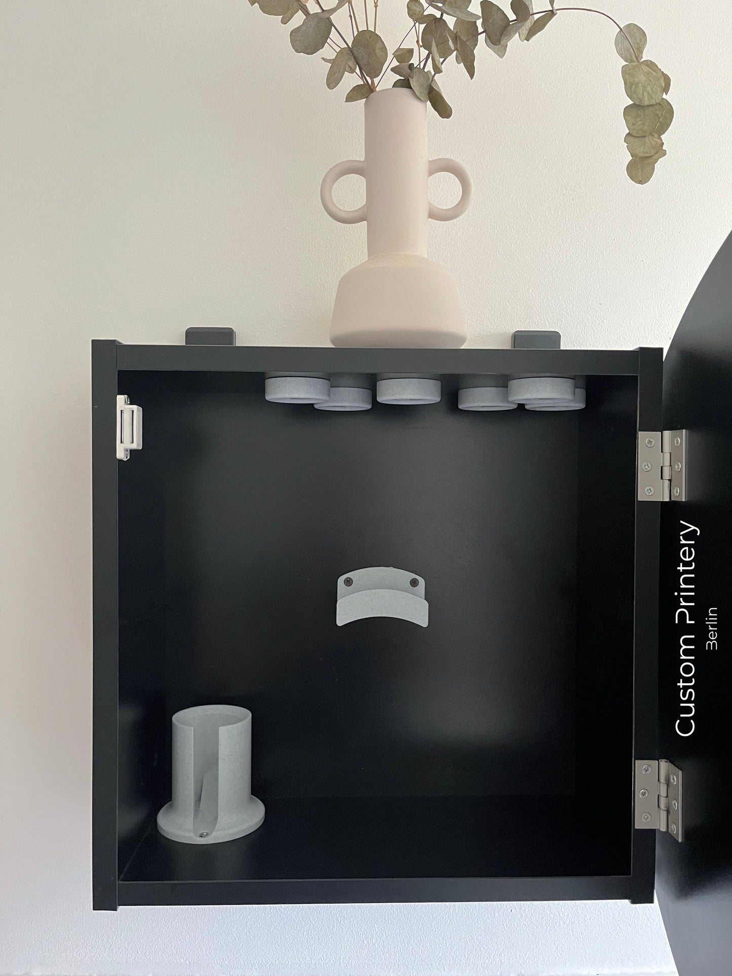 Schwarze Wandhalterung für Dyson Haarstyling-Gerät und Zubehör. Die Halterung enthält den Haartrockner, verschiedene Styling-Aufsätze und ist sauber organisiert. Oben auf dem Regal steht eine moderne Vase mit getrockneten Eukalyptuszweigen.