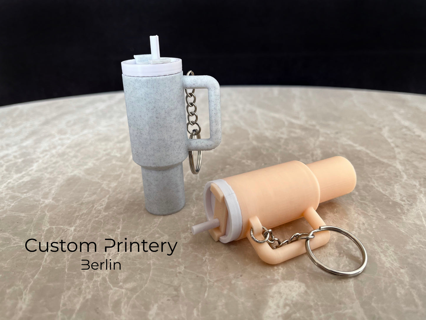 Mehrere bunte Schlüsselanhänger im Stil des Stanley Quencher Cups sind auf einer Marmoroberfläche arrangiert. Die Miniaturbecher haben Henkel, Trinkhalme und verschiedene Farben, darunter Schwarz, Blau, Pink, Rot, Gelb, Beige und Grau.