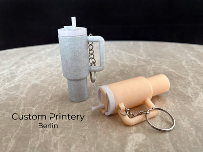 Mehrere bunte Schlüsselanhänger im Stil des Stanley Quencher Cups sind auf einer Marmoroberfläche arrangiert. Die Miniaturbecher haben Henkel, Trinkhalme und verschiedene Farben, darunter Schwarz, Blau, Pink, Rot, Gelb, Beige und Grau.