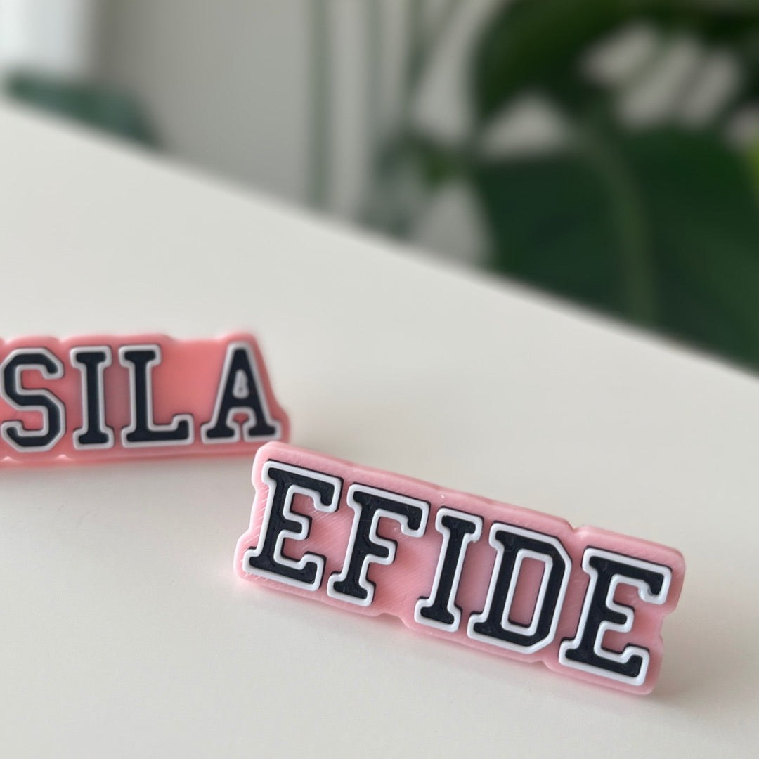 Custom black Crocs with colorful personalized name charms for 'Lena,' 'Noah,' 'Theo,' and 'Clara' from Custom Printery Berlin, showcased in a casual indoor setting with green plants.