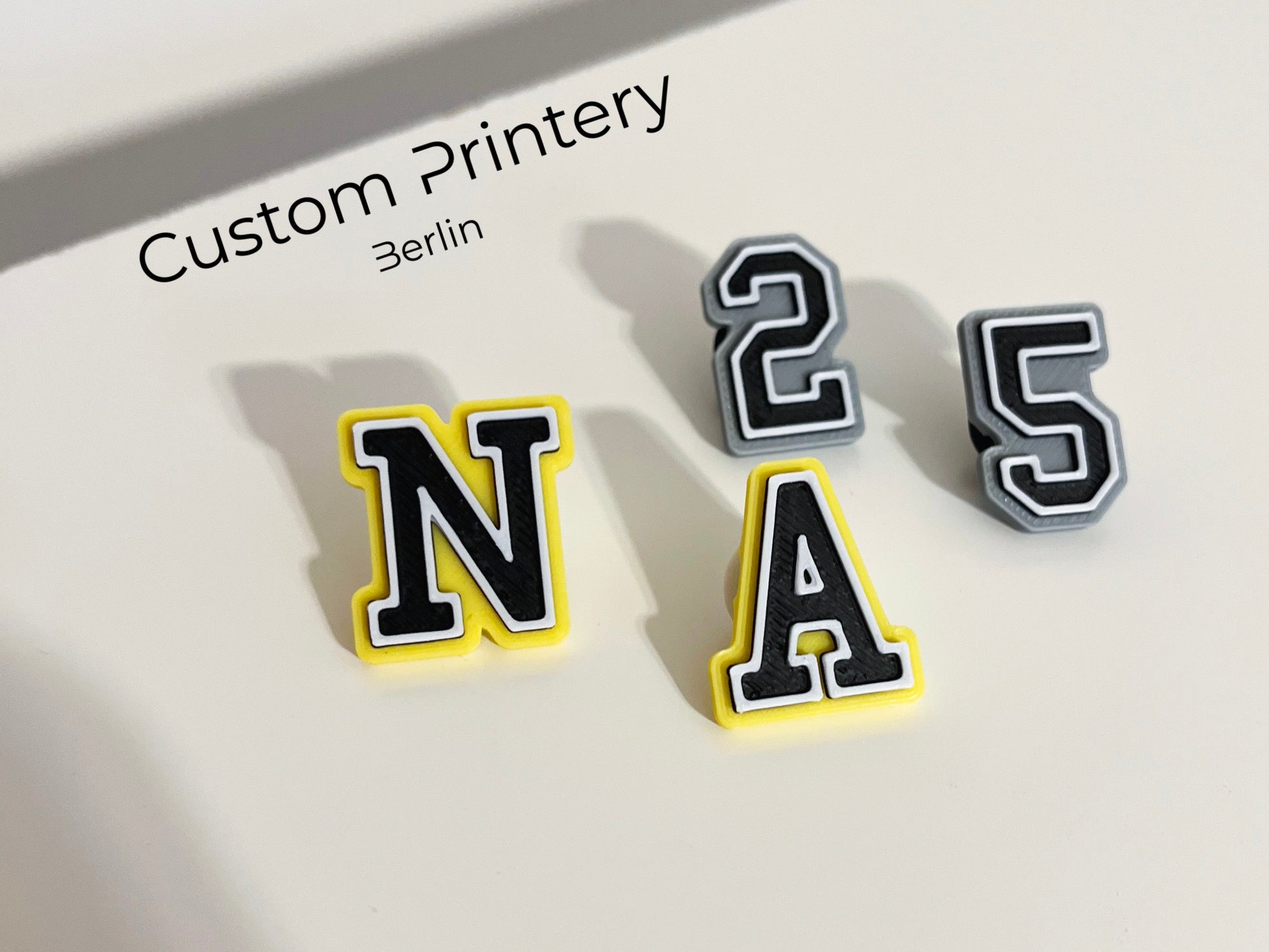 Custom black Crocs with colorful personalized name charms for 'Lena,' 'Noah,' 'Theo,' and 'Clara' from Custom Printery Berlin, showcased in a casual indoor setting with green plants.