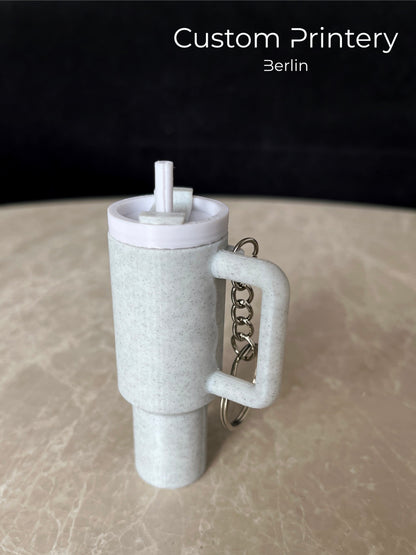 Mehrere bunte Schlüsselanhänger im Stil des Stanley Quencher Cups sind auf einer Marmoroberfläche arrangiert. Die Miniaturbecher haben Henkel, Trinkhalme und verschiedene Farben, darunter Schwarz, Blau, Pink, Rot, Gelb, Beige und Grau.