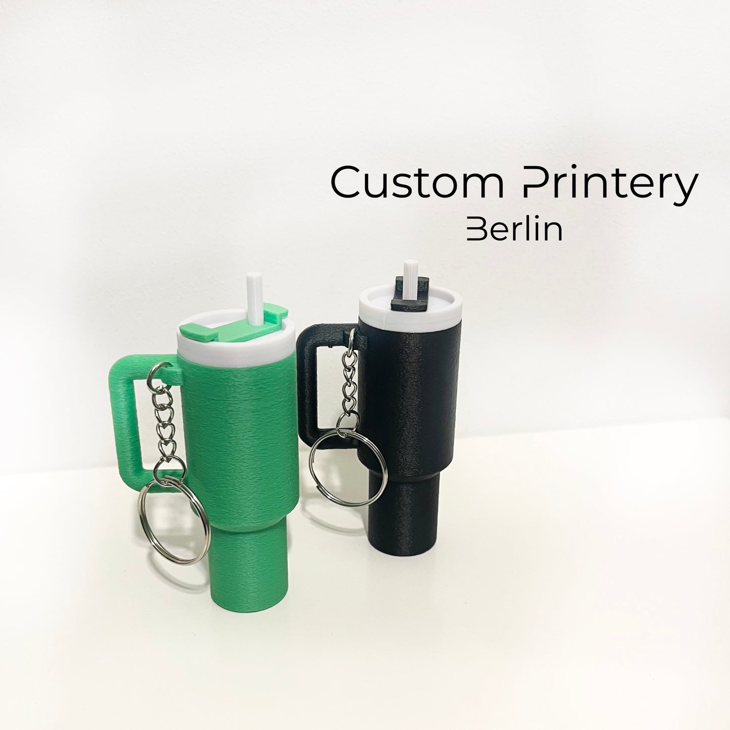 Mehrere bunte Schlüsselanhänger im Stil des Stanley Quencher Cups sind auf einer Marmoroberfläche arrangiert. Die Miniaturbecher haben Henkel, Trinkhalme und verschiedene Farben, darunter Schwarz, Blau, Pink, Rot, Gelb, Beige und Grau.