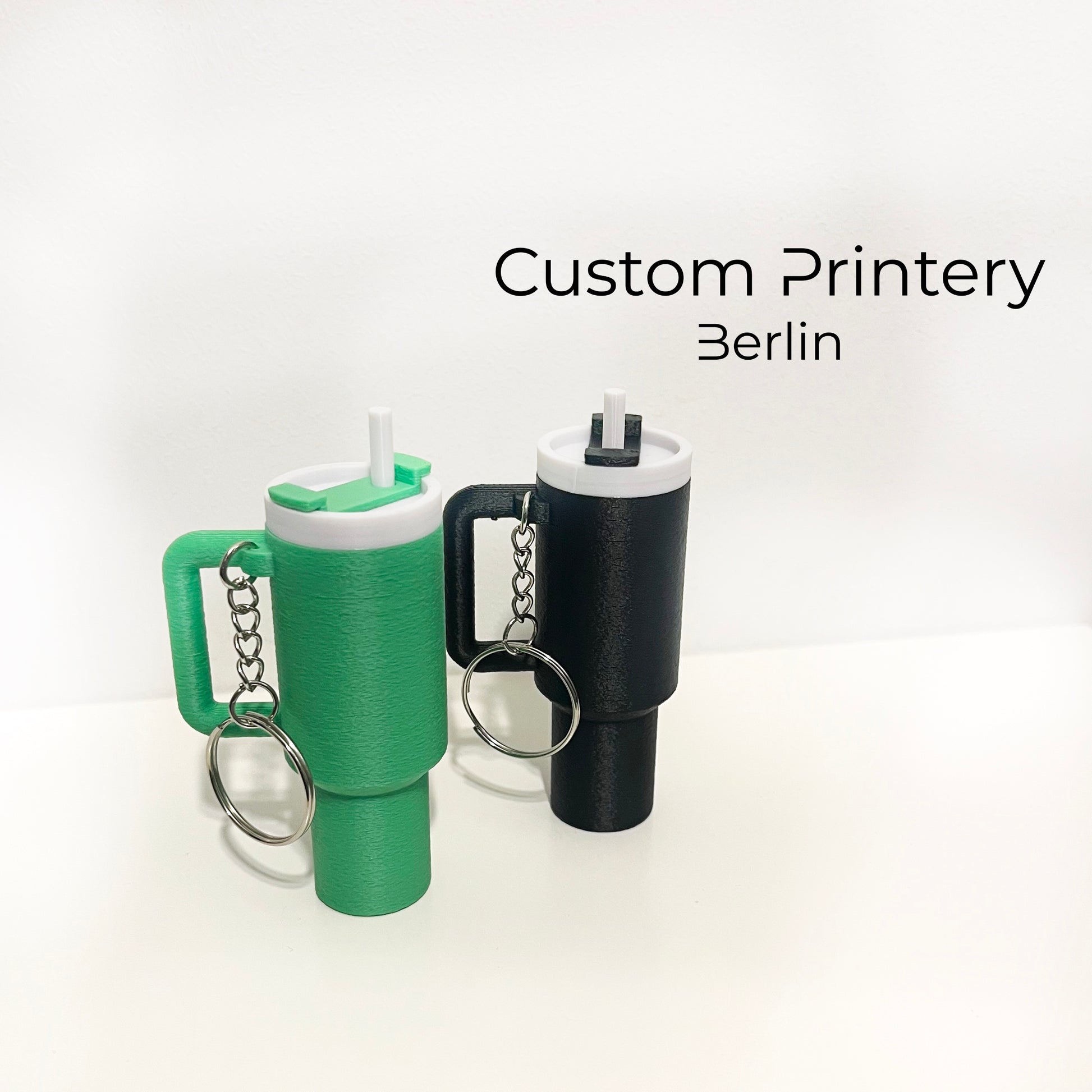 Mehrere bunte Schlüsselanhänger im Stil des Stanley Quencher Cups sind auf einer Marmoroberfläche arrangiert. Die Miniaturbecher haben Henkel, Trinkhalme und verschiedene Farben, darunter Schwarz, Blau, Pink, Rot, Gelb, Beige und Grau.