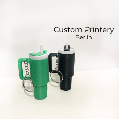 Mehrere bunte Schlüsselanhänger im Stil des Stanley Quencher Cups sind auf einer Marmoroberfläche arrangiert. Die Miniaturbecher haben Henkel, Trinkhalme und verschiedene Farben, darunter Schwarz, Blau, Pink, Rot, Gelb, Beige und Grau.