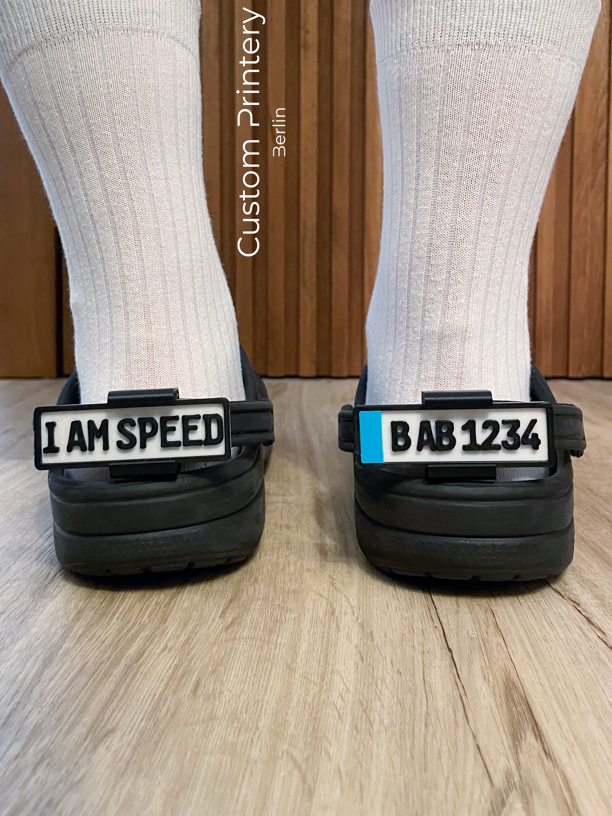 Custom black Crocs with colorful personalized name charms for 'Lena,' 'Noah,' 'Theo,' and 'Clara' from Custom Printery Berlin, showcased in a casual indoor setting with green plants.