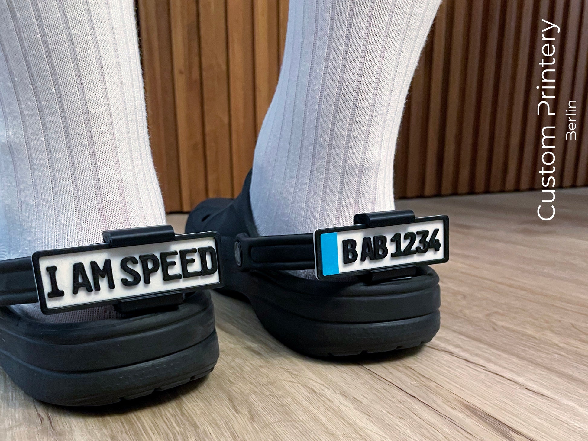 Custom black Crocs with colorful personalized name charms for 'Lena,' 'Noah,' 'Theo,' and 'Clara' from Custom Printery Berlin, showcased in a casual indoor setting with green plants.