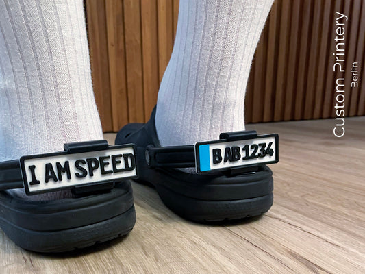 Custom black Crocs with colorful personalized name charms for 'Lena,' 'Noah,' 'Theo,' and 'Clara' from Custom Printery Berlin, showcased in a casual indoor setting with green plants.