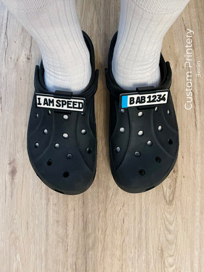 Custom black Crocs with colorful personalized name charms for 'Lena,' 'Noah,' 'Theo,' and 'Clara' from Custom Printery Berlin, showcased in a casual indoor setting with green plants.