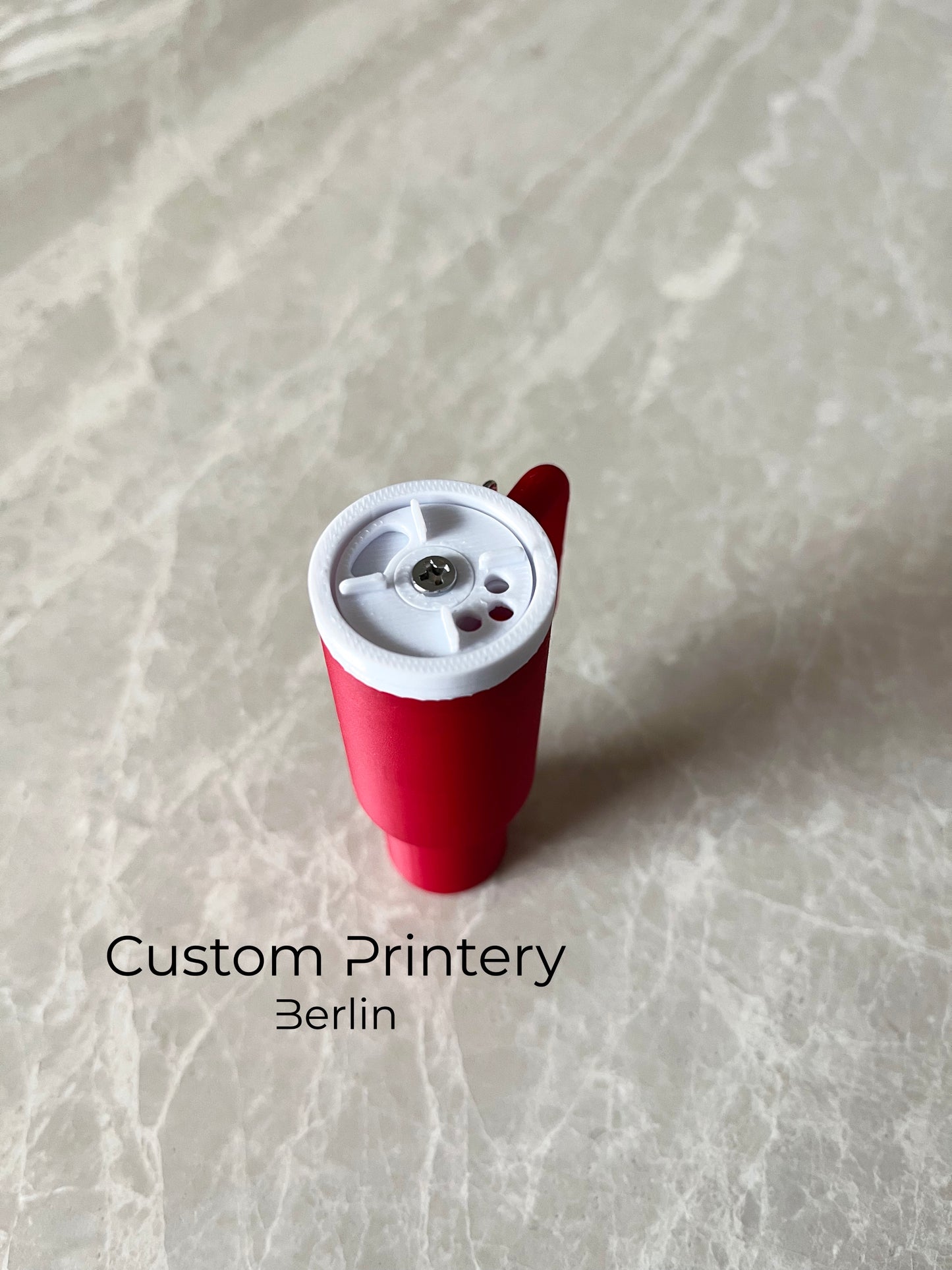 Mehrere bunte Schlüsselanhänger im Stil des Stanley Quencher Cups sind auf einer Marmoroberfläche arrangiert. Die Miniaturbecher haben Henkel, Trinkhalme und verschiedene Farben, darunter Schwarz, Blau, Pink, Rot, Gelb, Beige und Grau.