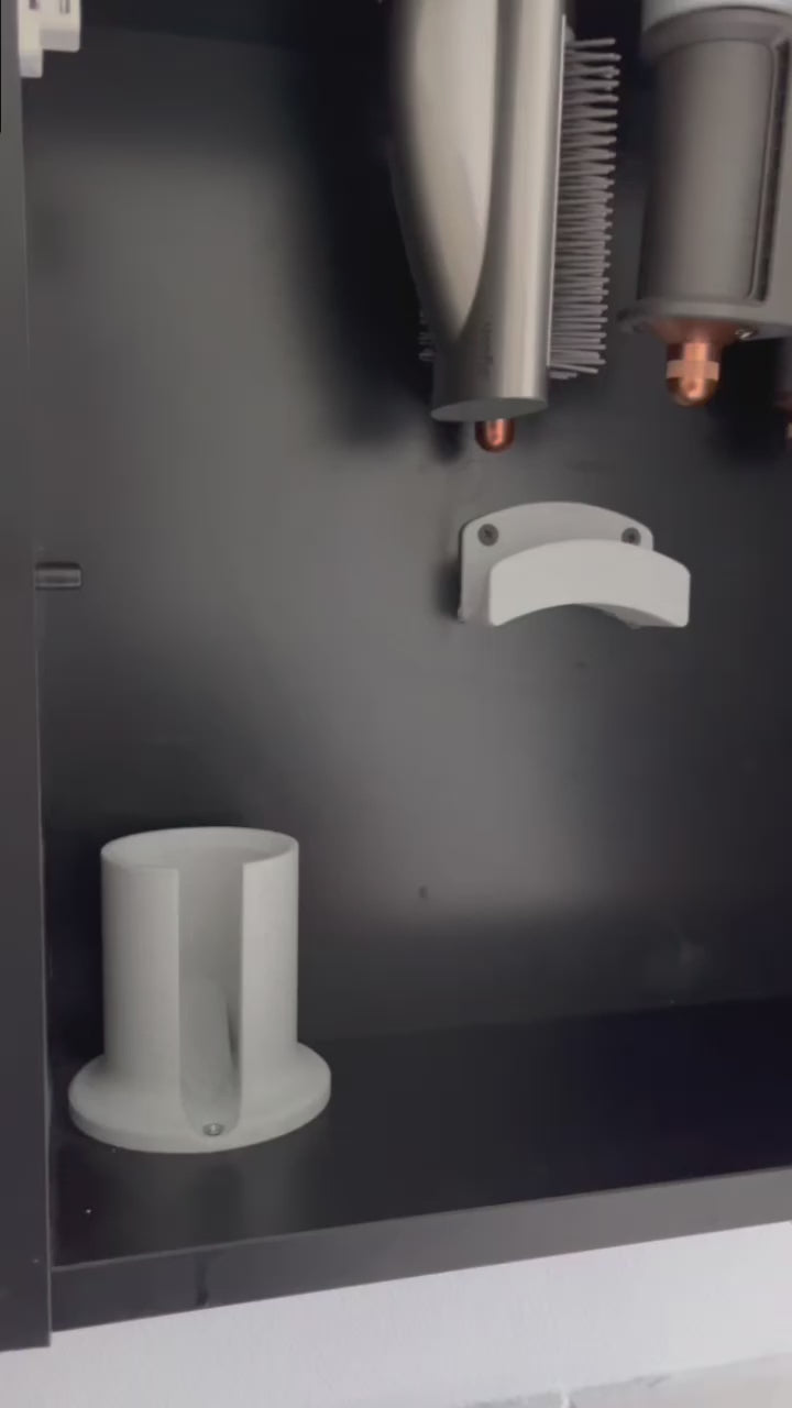 Schwarze Wandhalterung für Dyson Haarstyling-Gerät und Zubehör. Die Halterung enthält den Haartrockner, verschiedene Styling-Aufsätze und ist sauber organisiert. Oben auf dem Regal steht eine moderne Vase mit getrockneten Eukalyptuszweigen.